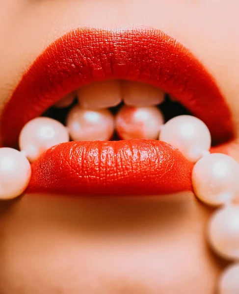 Sexy bright red woman lips holding pearl necklace close up photo. Female lips with luxury orange red lipstick. Woman holding collar of pearl in the mouth close up photo