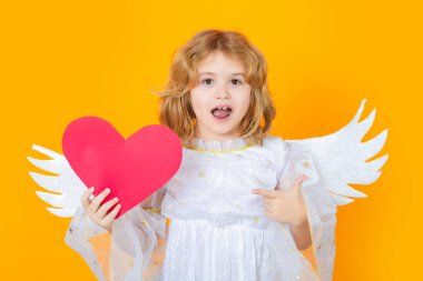 Child angel pointing on paper red heart, point gesture. Kid wearing angel costume white dress and feather wings. Innocent child. Little angel on isolated studio background