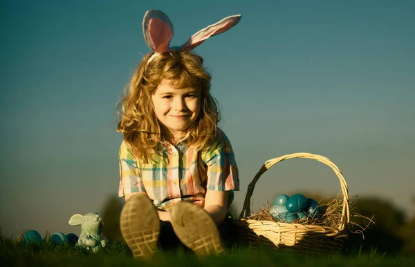 Cute bunny child boy with rabbit ears. Children hunting easter eggs on sky background with copy space. Child with easter eggs and bunny ears, outdoor portrait