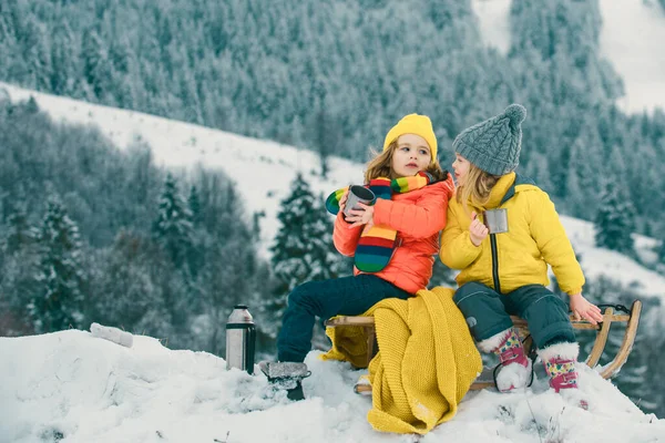 Winter camp picnic for children. Funny little boy and girl in winter clothes walks during a snowfall. Outdoors winter activities for kids. Cute children drink warm tea