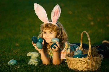 Paskalya yumurtalarını sepete koyan çocuk. Çocuk parkta çimlerin üzerinde yatıyor. Paskalya yumurtası avı. Fynny çocuk suratı. Tavşan kulaklı çocuk.