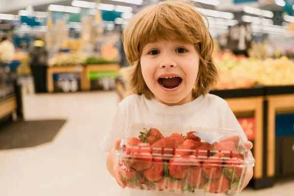 Cute toddler boy in a food store or a supermarket choosing fresh organic strawberry. Healthy lifestyle for young family with kids. Cute boy child with shopping trolley with products