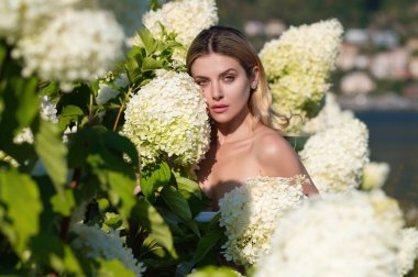 Spring girl in blooming bush of hydrangea flowers in spring garden. Hydrangeas flowers. Woman near a blossoming hydrangea in a garden. Sexy beautiful woman with naked shoulder in blooming garden