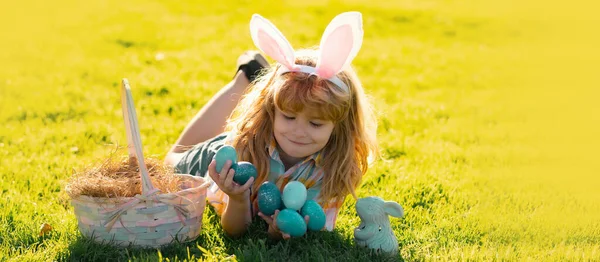 Child boy hunting easter eggs, laying on grass. Kid in rabbit bunny ears outdoor. Easter bunny children. Spring holiday. Panoramic web banner frame