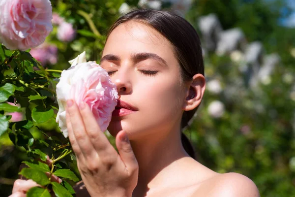 Beautiful girl smelling a rose flower in spring park. Young woman in flowering garden with roses. Beauty model with spring flowers. Happy spring. Pretty woman enjoying smell flowers in spring park