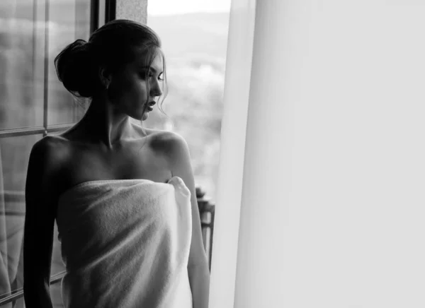 窓の近くにシャワーを浴びて外を見ると 官能的な若い女性がタオルに包まれました ウィンドウの背景に豪華なメイクと髪型を持つ美しい女性 — ストック写真