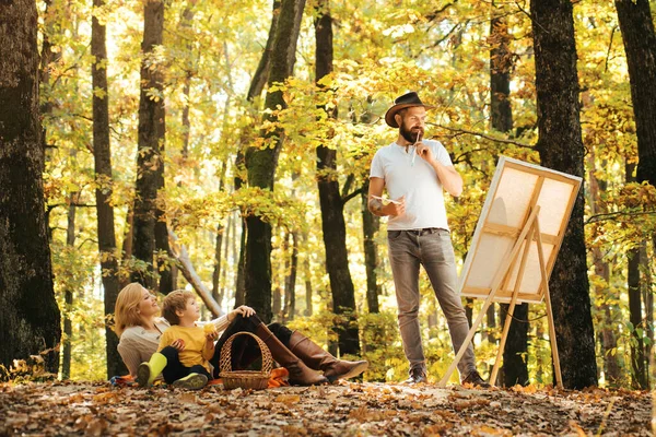 Start new picture. Portrait of young bearded smiling middle age white man artist drawing in open plein air outside with watercolor on paper at easel. Family picnic at autumn nature