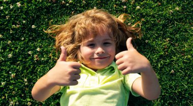 Bahar çocukları portresi olan pankart. Çimlerin üzerinde başparmak işareti olan komik çocuk, yüzünü kapat. Çocuklar yazın tadını çıkarıyor. Yeşil çim arka plandaki çocuklar