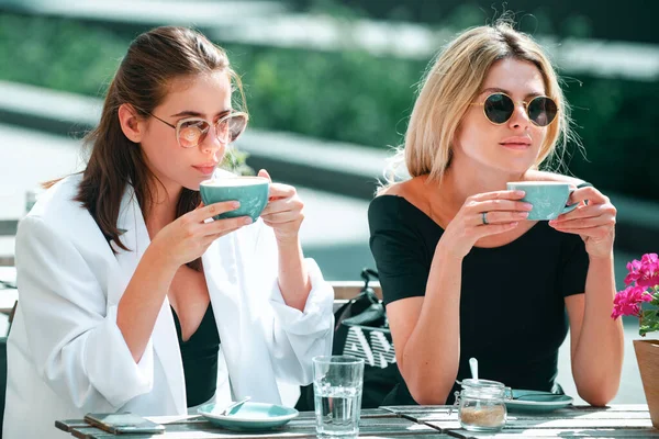 Girl friends in cafe outdoor. Two beautiful girls with cups coffee in summer cafe. Outdoors portrait of two young beautiful women friends drinking coffee. Beautiful girls metting in cafe