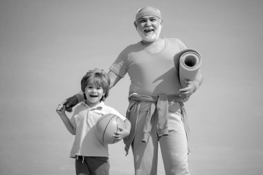 Büyükbabam ve torunum ellerinde basketbol topu ve yoga minderiyle. Kıdemli bir adam ve küçük tatlı bir çocuk mavi gökyüzü arka planında egzersiz yapıyorlar - izole edilmiş