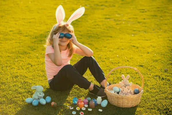 Child boy with bunny ears cover eyes with eggs on grass background