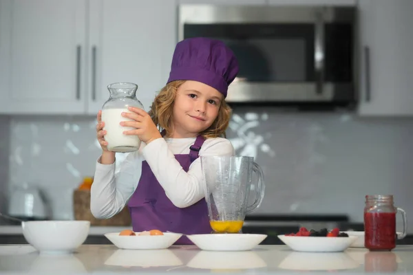 Child food preparation. Funny little kid chef cook hold bottle milk wearing uniform cook cap and apron cooked food in the kitchen