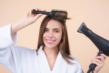 Bornozlu kadın saç tarıyor, saç kurutma makinesiyle saç kurutuyor. Saçlarını tarayan kadın modelin portresi. Saç fırçası ve saç kurutma makinesi olan kız. Saç bakımı ve güzellik. Sabah rutini