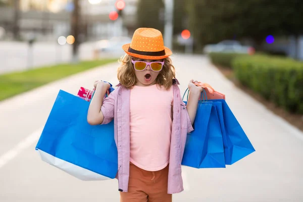 Surprized shopper. Excited kid in fashion clothes goes shopping. Amazed kid with shopping packages outdoor. Shopper child with shopping bag walking on street and carrying shopping bags