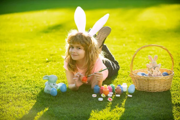 Easter kid in park. Easter kids boy in bunny ears paint easter eggs outdoor. Cute child in rabbit costume with bunny ears having fun and painting eggs in park