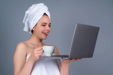Morning freelancer working on laptop. Business woman with towel on head with coffee working on laptop. Morning coffee. Portrait of smiling woman with cup of coffee using laptop in gray studio