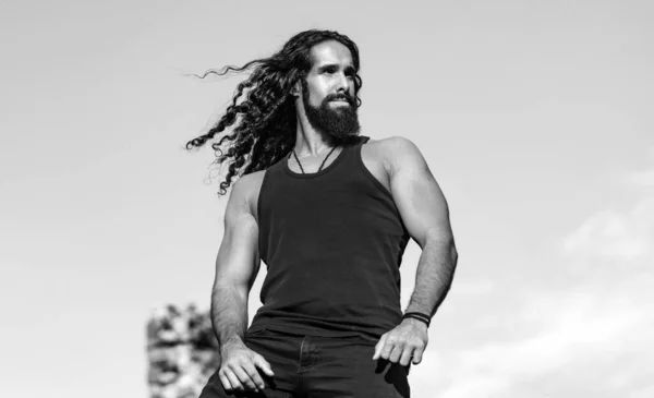 Handsome bearded man with long dark hair. Fashionable man with long wavy hair and strong healthy body. Mens style and wellness concept. Strong muscular handsome man at blue summer sky background