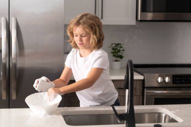 Çocuk mutfağın içinde bulaşık yıkıyor. Mutfak lavabosundaki süngerle bulaşık deterjanı. Çocuk anne ve babasına ev işlerinde yardım ediyor