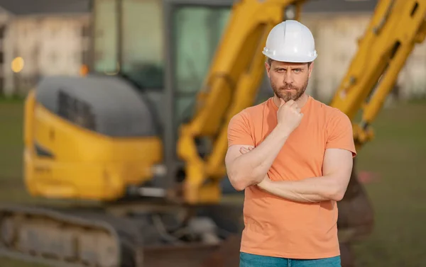 Builder near excavator. Builder worker with excavator. Builder in helmet. Worker in hardhat. Portrait mechanical worker in construction helmet. Engineer builder foreman or repairman