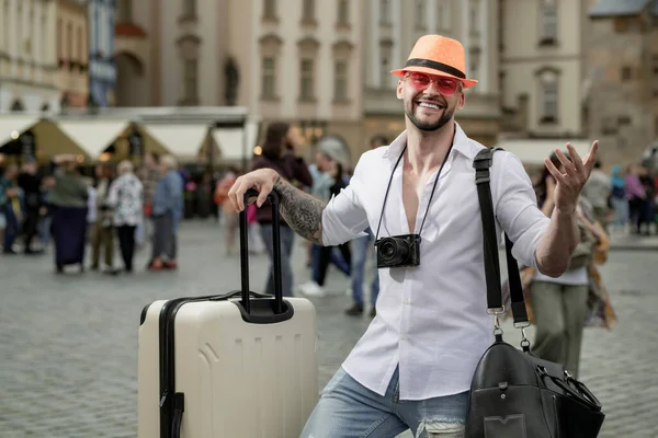 Stock image Traveller, fashion tourist. Tourist man in fashion hat jeans and sunglasses with suitcase walking on street. Man traveler with travel bag outdoor. Portrait of young caucasian man in hat and sunglasses