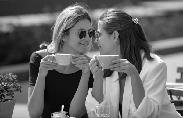 Two young women talk and drink coffee in cafe, outdoors. Young women with coffee cups at cafe. Europe vacation