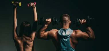 Dambılla egzersiz yapan seksi spor çifti. Çıplak vücutlu kaslı bir adam, arka planda koyu renkli halterleri olan fitness kadını. Sıska seksi kız güçlü kaslı adam egzersizi yapıyor.