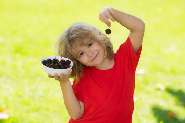 Cute little boy in red t-shirt eating cherries, making funny faces and playing with the cherries, having fun