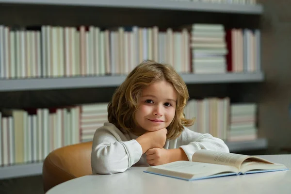 緊張した生徒 学校の図書館で机の上で宿題をしている少年 子供は本の山の近くで宿題文学を勉強する 子供は背景に本の棚のある本を読む — ストック写真