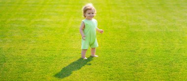 Baharda çim tarlasında bebek, afiş. Yeşil çimlerin üzerinde çıplak ayakla duran bebek. Küçük çocuk yaz parkında oynuyor. Mutlu çocukluk ve çocuk sağlığı hizmetleri