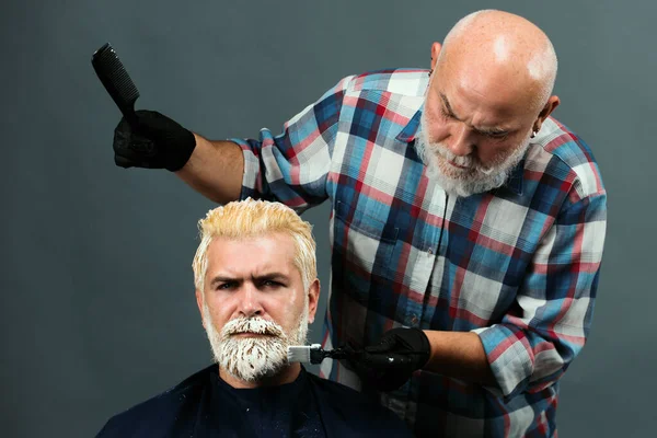 Bearded man coloring hair. Hair salon, hair coloring man. Attractive senior barber doing a haircut and haircolor for client at barber shop. Hipster guy dye his hair color on a gray background