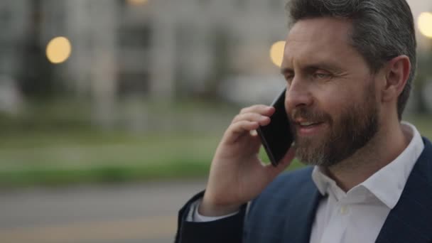 ビジネスマンは携帯電話を使って外で正式なスーツを着ていた 中年の男性が通りを歩いて電話や笑顔を話す 大人の男が電話で話してる 電話を使った正式な訴訟のビジネスマン — ストック動画