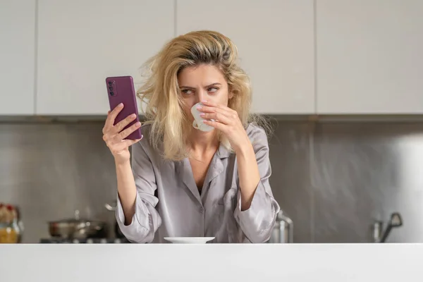 Upset woman reading message or bad new at smartphone in the kitchen. Sad woman wearing sexy pajama drink coffee in home kitchen
