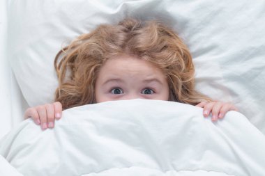 Yatakta saklanan korkmuş çocuk. Çocuk örtülü, yüzü battaniyeli. Çocuk yatakta yatıyor, uyanıyor. Sabah uyanışı, güneşli çocuklar için yatak odası. Kabus çocukları, beyaz battaniyenin altında saklanan çocuk, üst manzara.