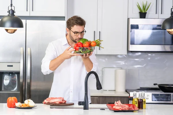 健康管理を行う 自宅のキッチンに立っているカジュアルなシャツのハンサムな男 台所で野菜を調理する若い男 サラダボウルで男を食事 グリーンサラダ健康食品のコンセプト — ストック写真
