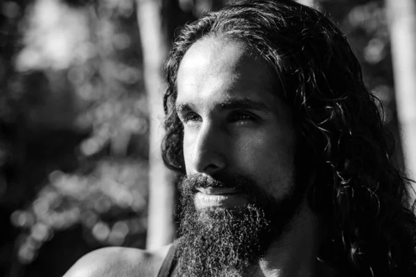 stock image Portrait of handsome young strong man with long dark hair looking away while standing at forest background. Sexy bearded model macho man. Man fashion and style concept