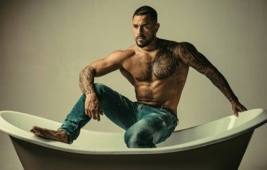 Banyodaki seksi maço adam. Şampanya şişesi tutan ve banyoda poz veren güçlü kaslı dövmeli bir adam. Banyoda seksi vücudu olan, kot pantolonlu, sakallı, gömleksiz yakışıklı adam.