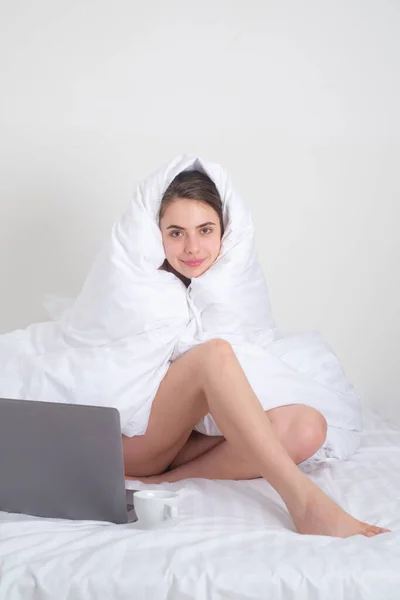 Woman watching laptop. Beautiful young woman covered with warm blanket using laptop on bed at home. Lazy freelancer girl with laptop sitting on bed. Girl work on laptop at morning resting in bed