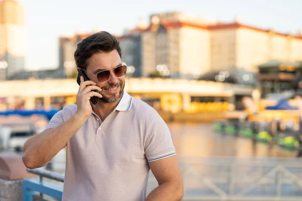 Portrait of handsome man chatting on phone outdoor. Stylish man talking on phone dressed in polo. Fashion male posing on the city street. Urban style. Business phone conversation