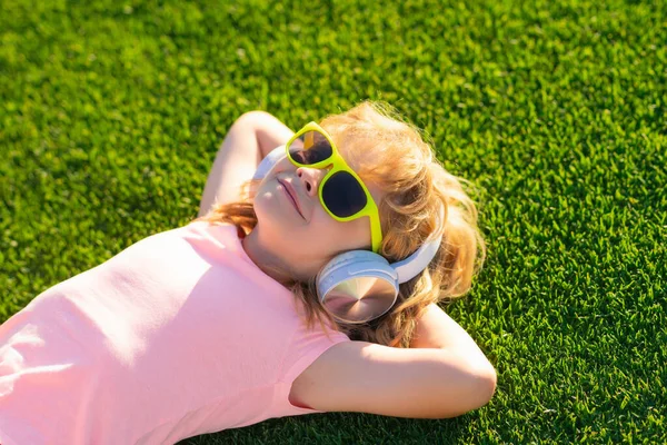 子供はヘッドフォンで音楽を聴くのが好きです 夢を見る子供は緑の草の背景の上のヘッドフォンで音楽を聞くのを楽しむ 夏の公園や裏庭の屋外で音楽を聴く — ストック写真