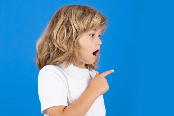 Surprised child looking away, pointing finger on isolated blue background. Shocked kid pointing to copyspace, showing promo offers, points away. Excited emotions, shock, omg and wow expression
