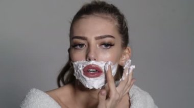 Yüzünde köpük olan kadın jileti elinde tutuyor. Meşgul yüzlü kız bornoz giyiyor, gri arka plan. Berber ve tıraş konsepti. Kadın trans, keskin jiletle cinsiyet değiştiriyor.