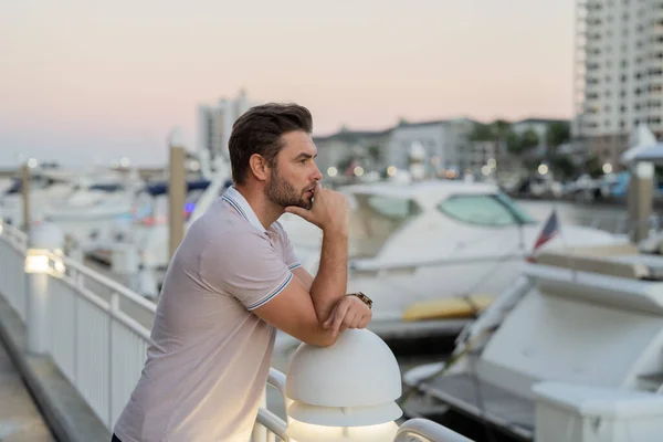 Rich businessman dreaming and thinking near the yacht. Portrait of confident man in modern big american city. Stylish lambersexual model. Sexy man dressed in polo