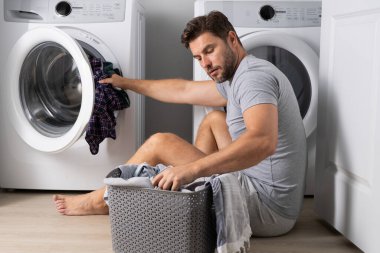 Çamaşır odasındaki çamaşır makinesine çamaşır yükleyen adam. Orta yaşlı bir adam çamaşır makinesine çamaşır koyuyor. Korkmuş ve ağzı açık bir şekilde hayret ediyor. Şaşırmak için.