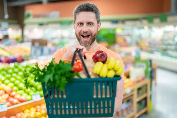 Man with shopping basket full of vegetables and fruits. Middle aged millennial man in a food store. Supermarket shopping and grocery shop concept. Man man 40s with shopping basket