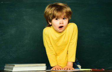 Okul malzemeleriyle masasının yanında gülen surat ifadesi olan sevimli bir çocuk. Okul çocukları yeşil karatahtaya karşı. Okul dersleri