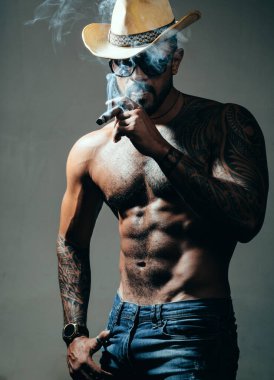 Sağlık. Sigara içiyor. duman üfler. kötü alışkanlık. güven karizması. dövme adam seksi abs. erkek modası. atletik vücutile kaslı maço adam. kovboy şapkalı acımasız sporcu