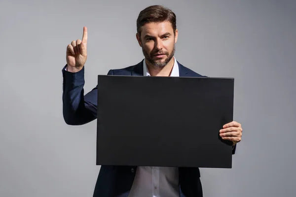 Business man in suit holding empty board and pointing index finger up on studio background. Guy showing blank signboard with copyspace. Idea and offer. Your advertisement. Blank advertising board