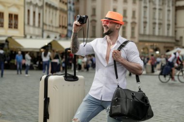 Moda şapkalı, güneş gözlüklü, bavullu, sokakta yürüyen bir turist. Seyahat çantasıyla seyahat eden bir adamın fotoğrafı. Çantalı adam seyahat, macera, tatil hayalleri kuruyor.