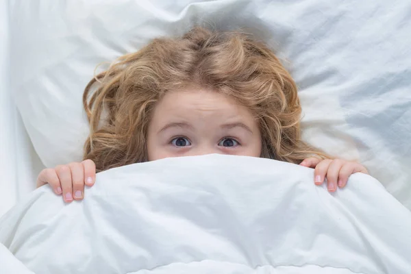 Nightmare kids, child hiding under white blanket, top view. Scared boy hiding in bed. Kid under covers, face cover with blanket. Morning awakening little child in bed. Kid wakes up in the bedroom