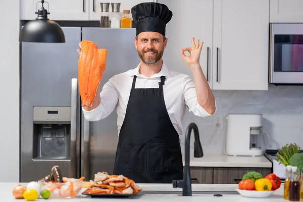 厨师围裙和厨师帽中的人在厨房准备新鲜的天然鲑鱼的肖像 一个漂亮而快乐的厨师正在准备生鲑鱼 健康食品 烹调概念 — 图库照片